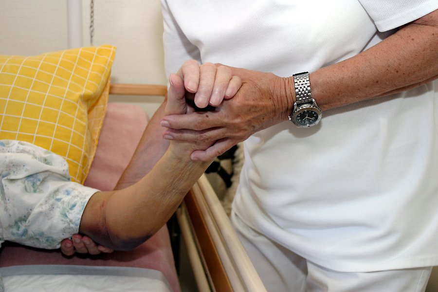 En person i sjukhuskläder håller i armen på en person som ligger i en säng.