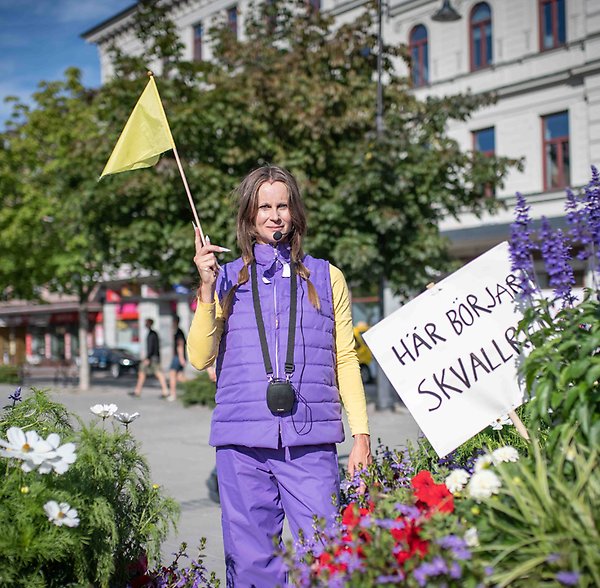 Konstnären Stina Nyberg står med flagga i handen vid skylten Här börjar Skvallret, korsningen Esplanaden, Sjögatan.