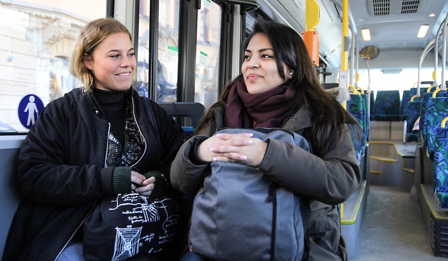 Två kvinnor sitter bredvid varandra i en buss med varsin väska i knät