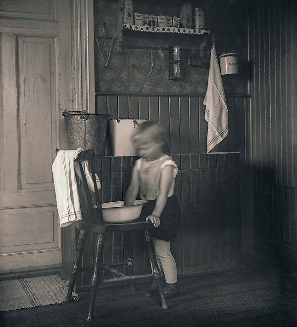 Ett barn står vid en stol och tvättar händerna i en balja i köksmiljö, bild.
