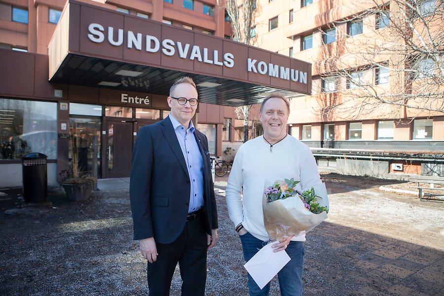 Två personer står framför en stor byggnad. Ovanför entrén står det Sundsvalls kommun med vita bokstäver. En av personerna håller i blommor.