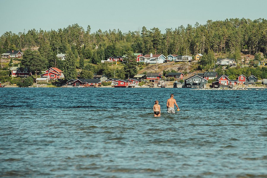 En vuxen och ett barn badar i havet utanför Sundsvall. Vattnet är blått, i bakgrunden ser man en ö med flera stugor på.
