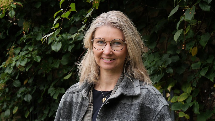 Ett porträttfoto av Linda Hallgren. Hon står framför ett träd iklädd en grå rock och har på sig ett par grå runda glasögon. 
