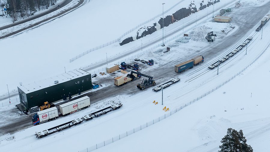Flygbild över snötäckt landskap där lastbilar och truckar rör sig på ett logistikområde med väg och järnvägsräls