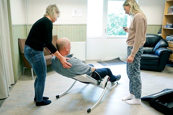 Två personer använder en elektrisk lyftstol för att lyfta en man som ligger ner på golvet.