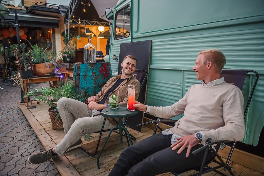Två män sitter på brassestolar på en uteservering på en restaurang. I handen har de varsin färgglad drink. De är glada och pratar med varandra.