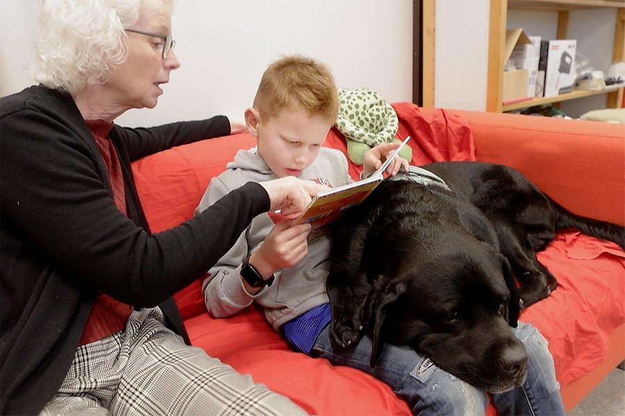 En kvinna och ett barn sitter med en hund i soffan och läser bok.
